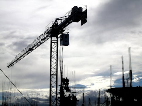 Silhouetted construction crane and workers against a cloudy sky during the day.
